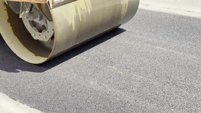 A yellow heavy road roller is seen working on a road construction and repair site, compacting a fresh layer of dark grey asphalt. The image showcases the construction equipment, steel drums, and the