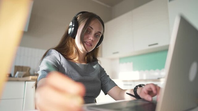 Young woman wears black headphone device. Happy woman types on silver laptop. Student works online from bright home kitchen. She smiles while using laptop screen. Freelance work at home desktop.
