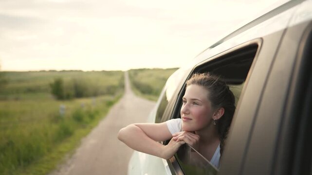 Happy girl leans out car window. Teenager enjoys travel along country road. Warm wind blows her braided hair on drive. She smiles from vehicle window. Nature surrounds happy road trip journey.