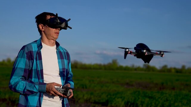 Drone pilot flies FPV drone in green field. Man wearing FPV goggles holds remote controller. Quadcopter hovers near pilot outdoors. FPV flying hobby at sunset. Drone operator controls flight.