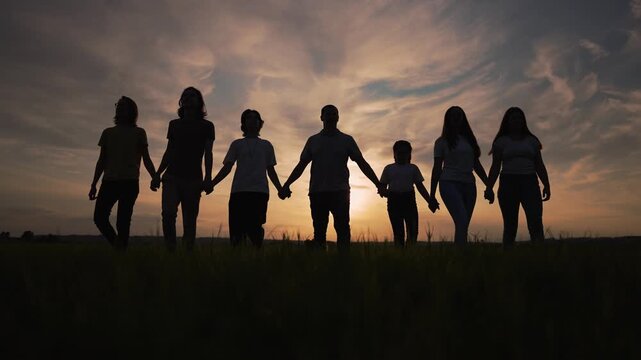 Large family group walking forward. Person holding hand in summer grass. Beautiful sunset forms dark silhouette. Group enjoys peaceful nature walking. Family bonding shows true togetherness.