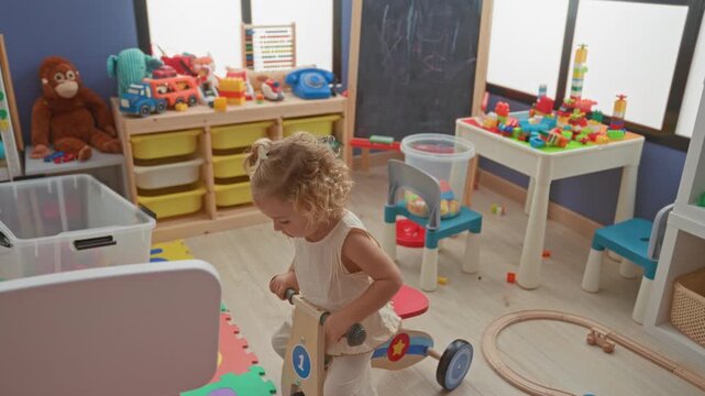 Little blonde girl riding a wooden tricycle holding the handlebars on a colorful number mat inside a building playroom with toy shelves and blocks; innocent curiosity.