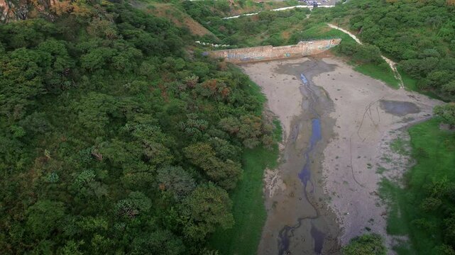 Abandoned dam with gang graffiti in dry riverbed Queretaro aerial view with green trees surrounding structure 4K drone POV public works infrastructure street art urban decay environmental scene