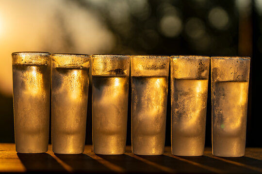Six ice-cold, frosted shot glasses lined up on wooden table, illuminated by the warm golden light of setting sun, closeup. Ideal for nightlife, party, summer bar, and celebration concepts