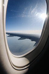 Fototapeta premium Airplane wing is visible through a passenger window above the clouds. Bright sun shines over the aircraft during a daytime flight and air travel journey background.