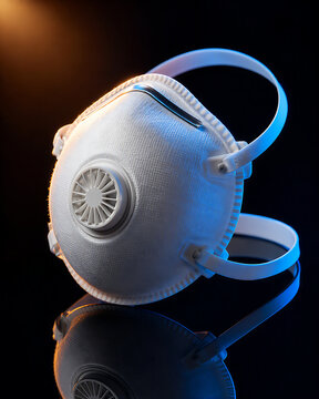 White N95 FFP2 respirator face mask with exhalation valve and elastic straps on dark reflective surface with blue and orange studio lighting, respiratory protection PPE for industrial safety content