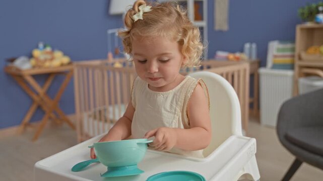 Little blonde girl holds a turquoise suction bowl and lifts its lid while sitting in a highchair inside building; curiosity.