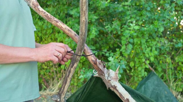 Close-up of a man hands securing wooden branch poles together with a plastic cable tie to build a wilderness bushcraft shelter and tarp setup for camping.