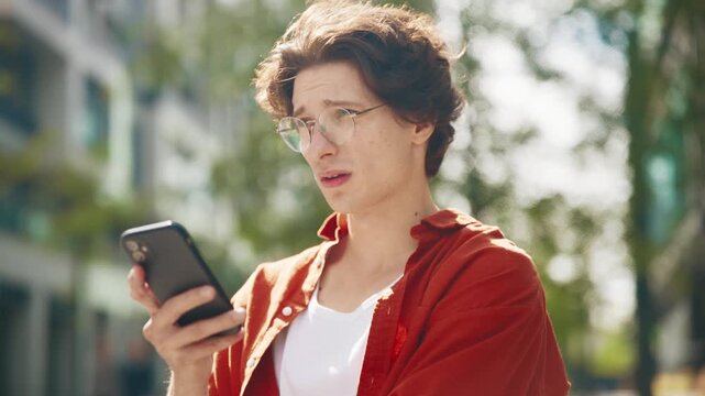 Man standing outdoors holding smartphone with thoughtful face. Wearing glasses and orange shirt looking away lost in thought. Reflecting during difficult chat in the street.