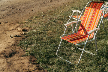 A colorful striped folding lawn chair stands on green grass next to a dusty dirt ground in bright daylight. © Dmitry