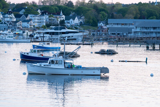 Boothbay Harbor, ME - May 27 2023 -View of moored boats in harbor and New England town in background with green trees. It is a cool spring morning.