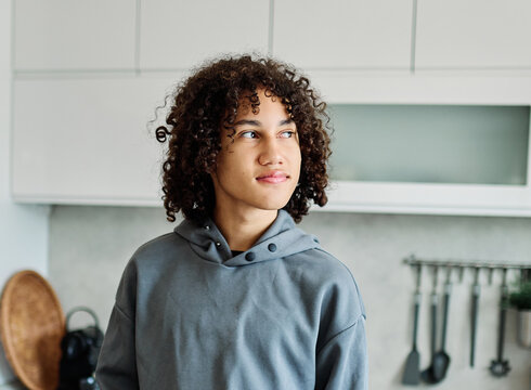 A thoughtful teen with curly hair stands in a modern kitchen wearing a gray hoodie, preparing to cook. Natural light and casual styling create a relaxed lifestyle portrait that conveys youthfulness, c