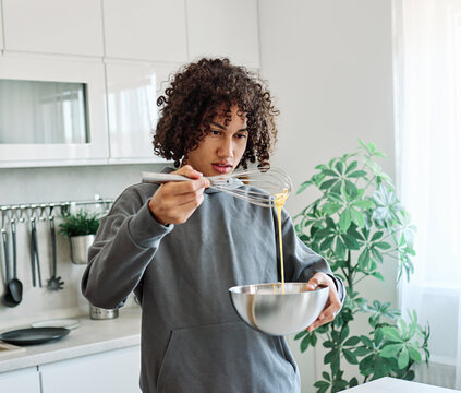 A teen concentrates while whisking eggs in a bright modern kitchen, practicing home cooking and breakfast preparation. The scene conveys focus, learning and independence, with natural light, plants an