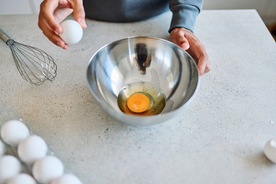 A teen cracks an egg into a stainless steel mixing bowl on a kitchen countertop, preparing to whisk for baking or breakfast. Visible hands, a whisk and an egg carton suggest home cooking, learning and