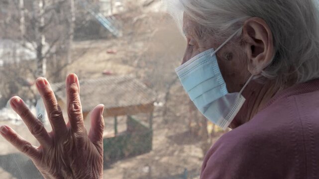 Solitary senior woman wearing a medical mask looking out window with hand on glass, feeling isolated and lonely while staying at home during global pandemic and quarantine