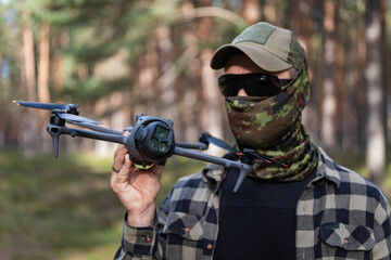 Fototapeta premium Professional tactical drone operator wearing a camouflage face mask and sunglasses holding a modern quadcopter with an advanced camera system in a sunny pine forest.