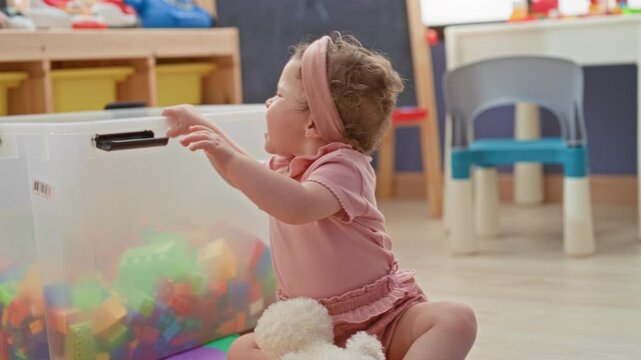 Toddler girl holding yellow building block and reaching toward a clear plastic toy bin on foam mat in playroom; curious discovery.