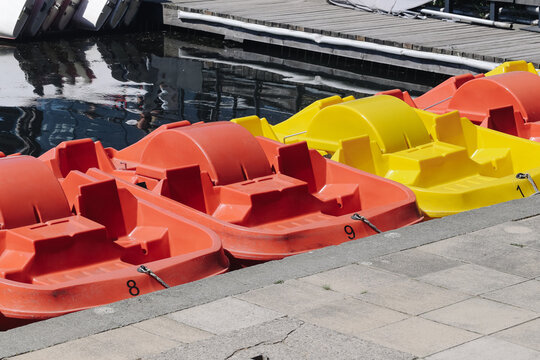 Red and yellow pedal boats are moored beside a paved lakeside dock. Colorful recreational boats wait at the water edge for summer leisure rides tourist rental and family fun background.