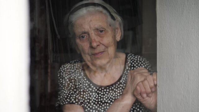 Portrait lonely old woman with grey hair and deep wrinkles looking sadly out window, hoping and waiting for someone to visit her in nursing home or during quarantine