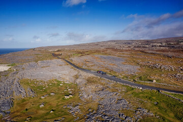 Winding road cuts through rugged limestone cliffs under a clear blue sky. Grassy patches and scattered rocks dominate the landscape. The scene feels remote and peaceful. Travel Ireland theme. © mark_gusev