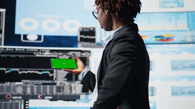 Manager compares company metrics on a green screen smartphone beside a digital video wall, reviewing KPIs before making business decisions.