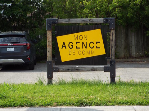 Gatineau, Quebec, Canada &ndash; July 02, 2026: A wooden framed outdoor business sign displays a yellow and black logo for "MON AGENCE DE COMM" near a parked vehicle" on boul. Saint-Ren&eacute; Ouest.