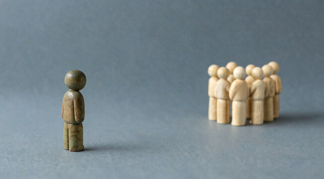 A single wooden figure stands apart from a group of figures huddled together. The scene evokes a sense of isolation and separation