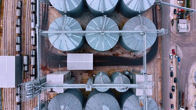 Expansive grain elevator complex situated alongside railways with tanker cars and roads with parked automobiles. Top view on the closely clustered storage tanks at the modern industrial plant.