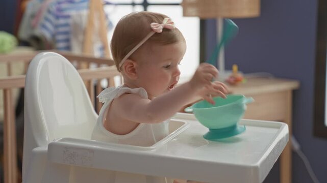 Girl holding spoon and turquoise suction bowl with bare arm in nursery highchair, exploring utensils and play eating at a wooden dresser table; curiosity.