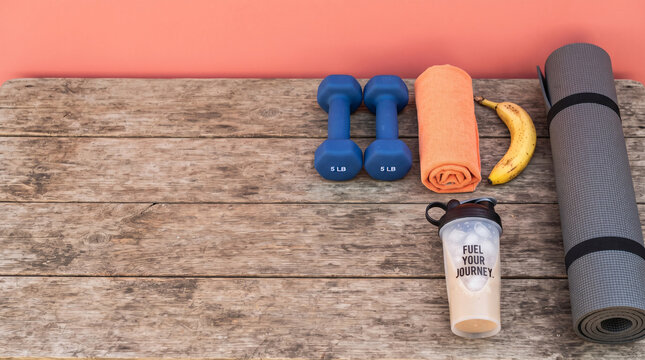 Fitness equipment collection on rustic wooden surface including blue dumbbells orange towel protein shaker banana and yoga mat over coral.