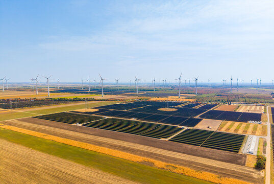 Large solar power plant surrounded by farmland and wind turbines. Aerial view of structured photovoltaic panel rows agricultural fields power lines and clear sky.