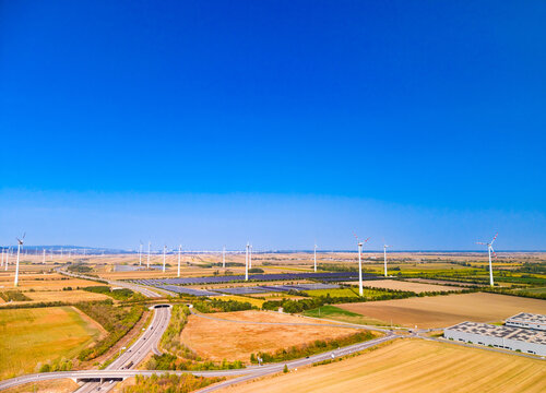 Aerial view of road highway and renewable energy facilities in rural landscape. A multi lane highway passes near solar panel fields and wind turbines under a clear sky.