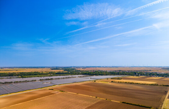 Wide aerial view of agricultural fields and distant solar installation. Plowed soil stretches toward a far solar array under a clear blue afternoon sky.