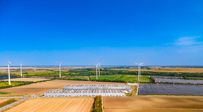 Aerial view of warehouse roof solar panels and wind turbines. Modern industrial structures stand among harvested fields beneath a clear blue sky.