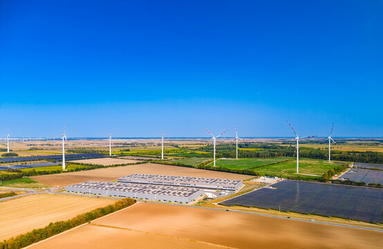 High angle view of renewable energy park and industrial facilities. Commercial buildings and photovoltaic arrays stretch across rural land under blue sky.