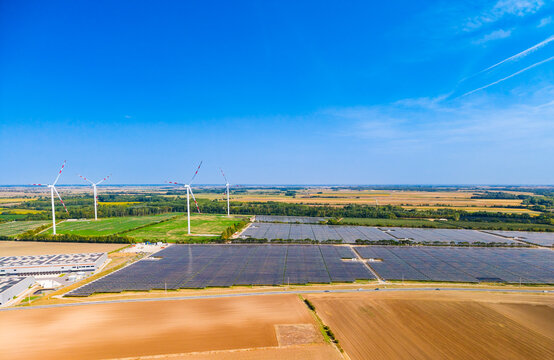 Aerial landscape of solar power station and wind farm. A thin condensation trail marks the bright blue sky above brown fields and collectors.