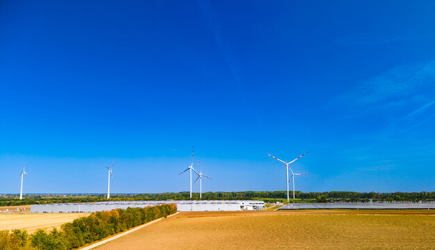 Wind turbines standing near industrial building in agricultural field. Eye level aerial view of wind turbines near warehouse facility surrounded by green and yellow fields