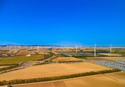 Wind turbine farm, wind generators across agricultural fields under sky. Wide high angle view of wind turbines and solar panels spread across farmland near highway under clear sky