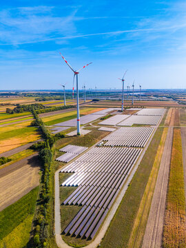 Wind turbine blades, green power generation on solar field. Vertical drone view of white wind turbine towering above long rows of ground mounted solar panels