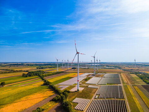 Wind generators and solar panel array in agricultural area. High angle shot of wind turbines, solar panel rows, agricultural landscape, blue sky with clear bright horizon.
