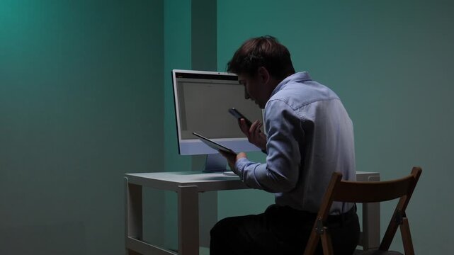 Man working late in a dim office, concentrating on multiple tasks, holding a tablet while talking on the phone and using a desktop computer, representing productivity and business communication