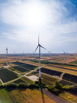 Elevated view of central wind turbine and solar park. Bright sunlight shines down on white generator blades above systematic silicon collectors and farmland.