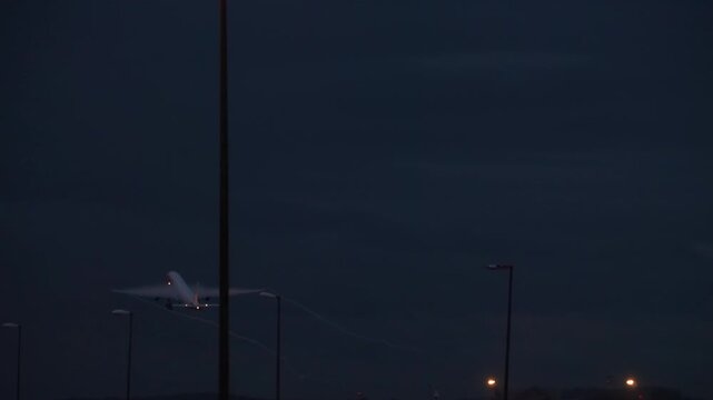Passenger jet taking off and climbing into dark night sky with flashing navigation lights and wingtip condensation trails, telephoto shot from airport airfield.