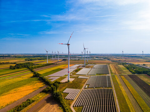 Renewable energy park with wind turbines and solar panels. Wide aerial view of wind turbines, rows of solar panels, colorful agricultural fields, blue sky on sunny day.