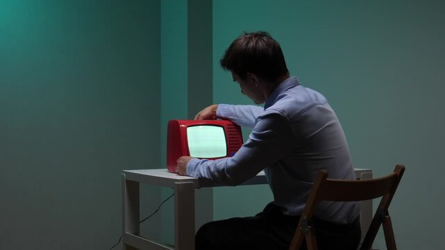 Man sitting at a desk in a dimly lit room, intensely focused on an old fashioned red television set displaying a blank, grainy screen, reflecting concepts of media consumption and vintage technology