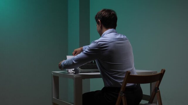 Man in a blue shirt sitting at a desk, typing on an old typewriter while throwing crumpled papers into a full trash can, symbolizing writer's block and creative struggles