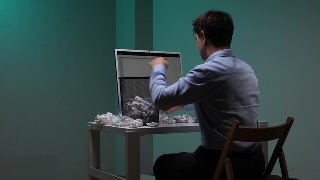 Man at desk overwhelmed by creative block and stress, tossing crumpled drafts into a small trash bin beside his computer during a frustrating work session