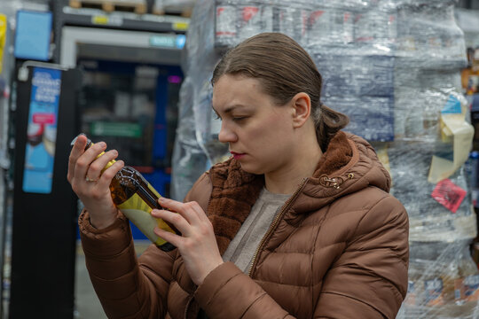 White woman scrutinizing beer ingredients and calories on label, concerned expression, holding bottle close, background pallets and shelving, sterile fluorescent lighting, focus on nutrition