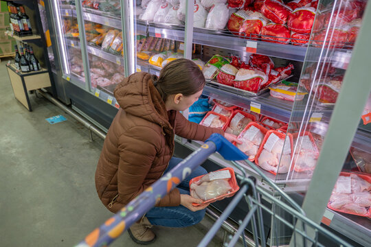 Woman closely inspecting frozen chicken package, reading nutrition facts and expiry date in supermarket fridge, casual jacket and cart visible, attentive shopper comparing quality and price for weekly