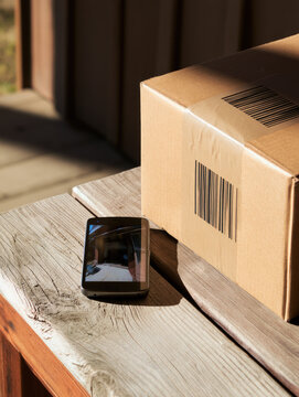 Cardboard shipping carton beside smart phone on wooden porch bench in warm sunlight, suggesting package delivery and home security monitoring with empty space for copy